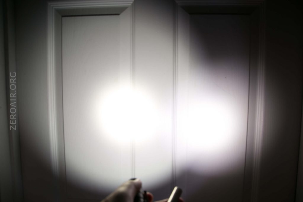 A white door is illuminated by two overlapping circles of light from flashlights, with a hand holding one flashlight visible at the bottom. The text ZEROAIR.ORG appears vertically on the left side.