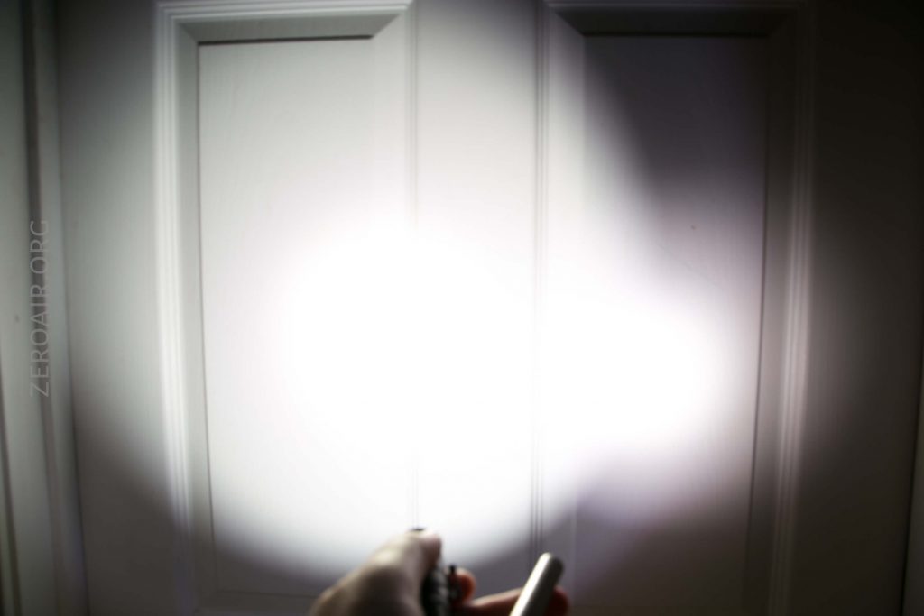 A person shines a flashlight at a white paneled door, creating a bright circular glare in the center of the door with some shadows around the edges.