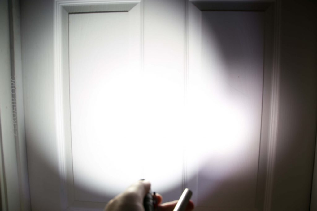 A hand holding a flashlight shines a bright beam of light onto a closed white paneled door, creating a large illuminated spot in the center of the door.