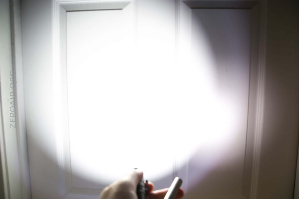A hand holding a flashlight points directly at a white door, creating a bright, circular beam of light on the door’s surface. The room appears otherwise dark.