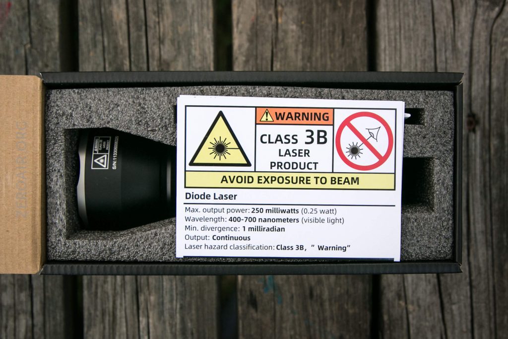 A black laser device in a foam case with a warning label indicating it is a Class 3B laser product. The label warns to avoid beam exposure and lists technical specifications and safety information.