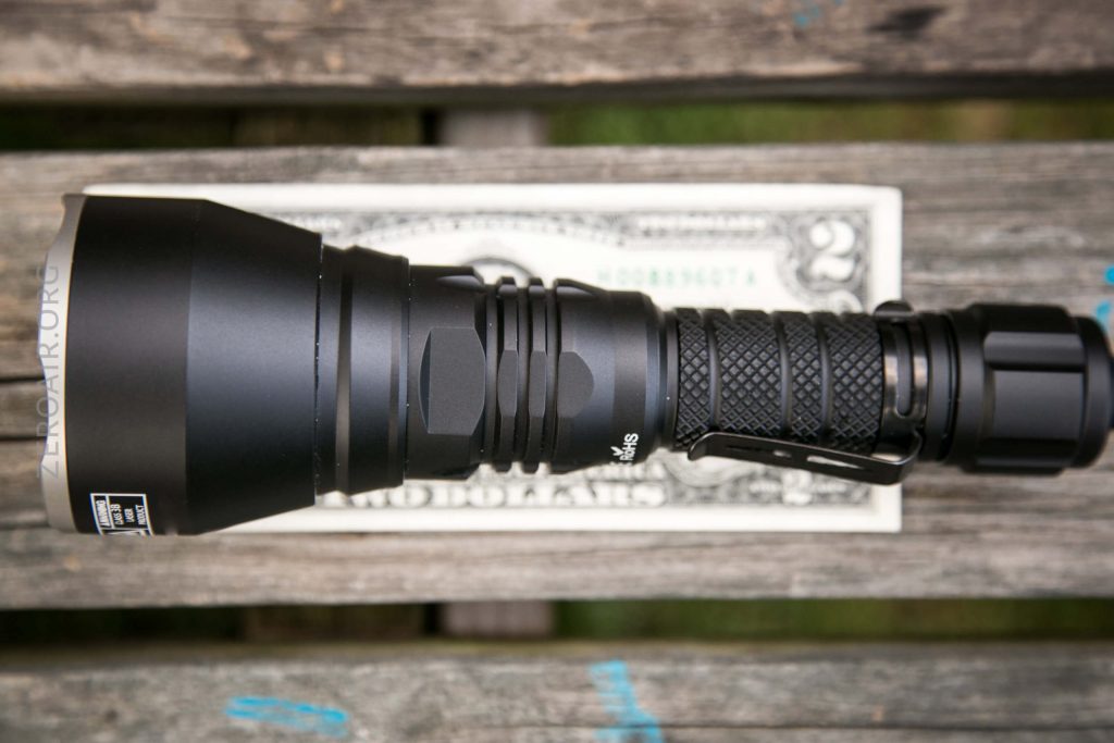 A black flashlight is placed horizontally on top of a U.S. two-dollar bill, showing the size comparison. The items rest on a weathered wooden surface.