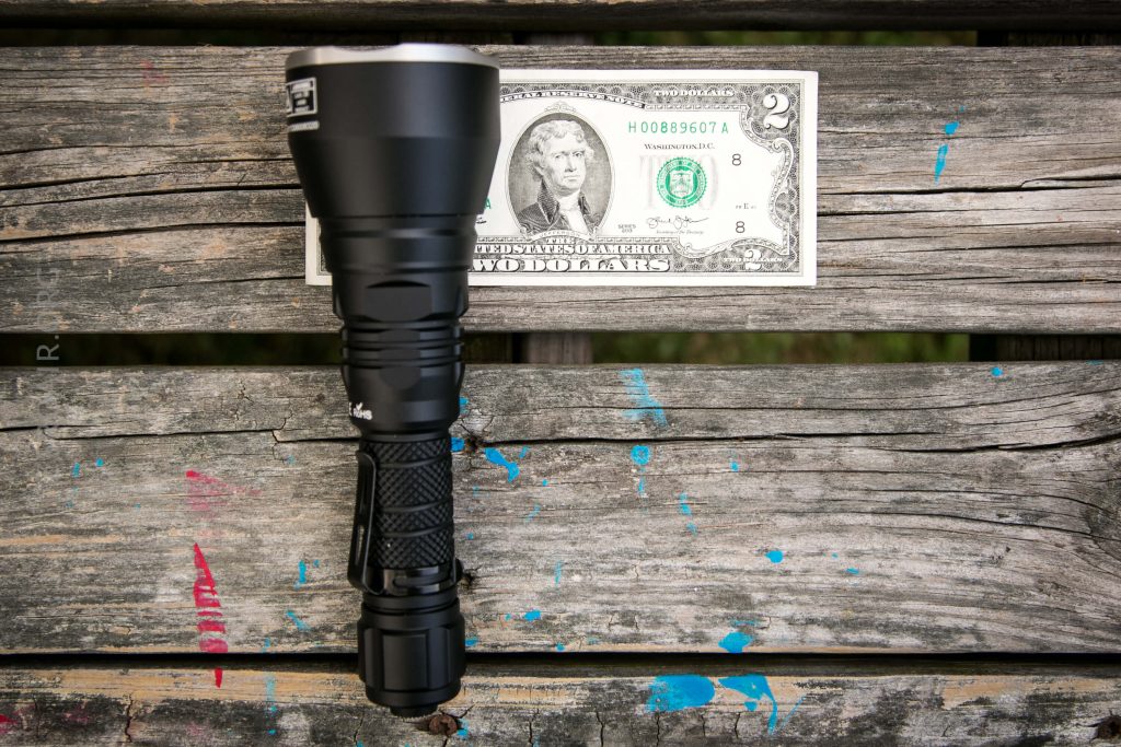 A black flashlight is placed horizontally next to a two-dollar bill on a weathered wooden bench with blue and red paint marks. The items are used to show a size comparison.