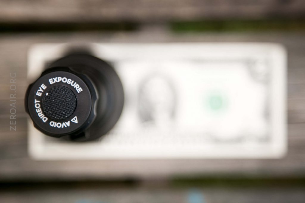 A close-up, top-down view of a flashlight head with “AVOID DIRECT EYE EXPOSURE” written on it, placed above a blurred US one-dollar bill on a wooden surface.