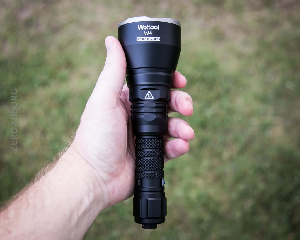 A hand holding a black Weltool W4 Dragon in Cloud flashlight outdoors, with grass in the background and ZEROAIR.ORG faintly visible on the left side of the image.