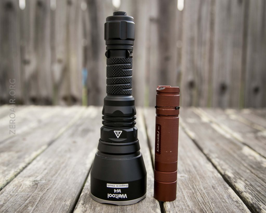 Two flashlights stand upright on a wooden surface, one large black flashlight on the left and a smaller, slimmer brown flashlight on the right. A wooden fence is visible in the background.