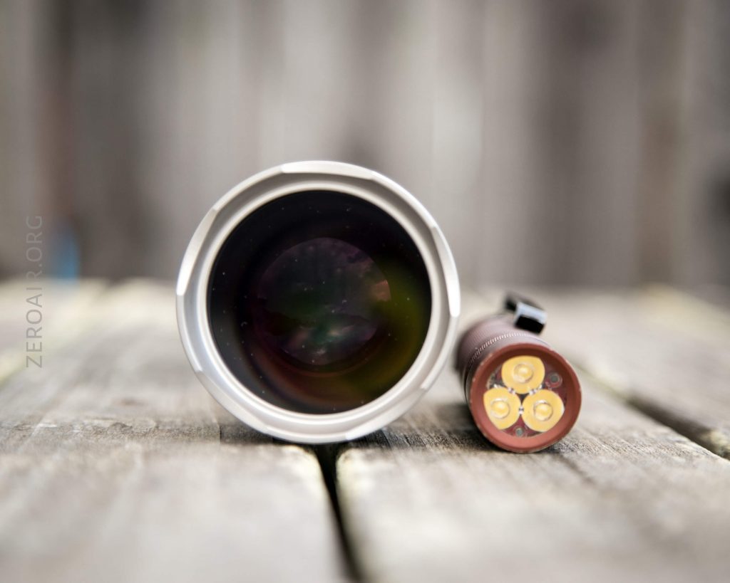 A close-up view of two cylindrical objects placed horizontally on a wooden surface. The object on the left shows a large glass lens, while the object on the right displays three yellow circles on its end.