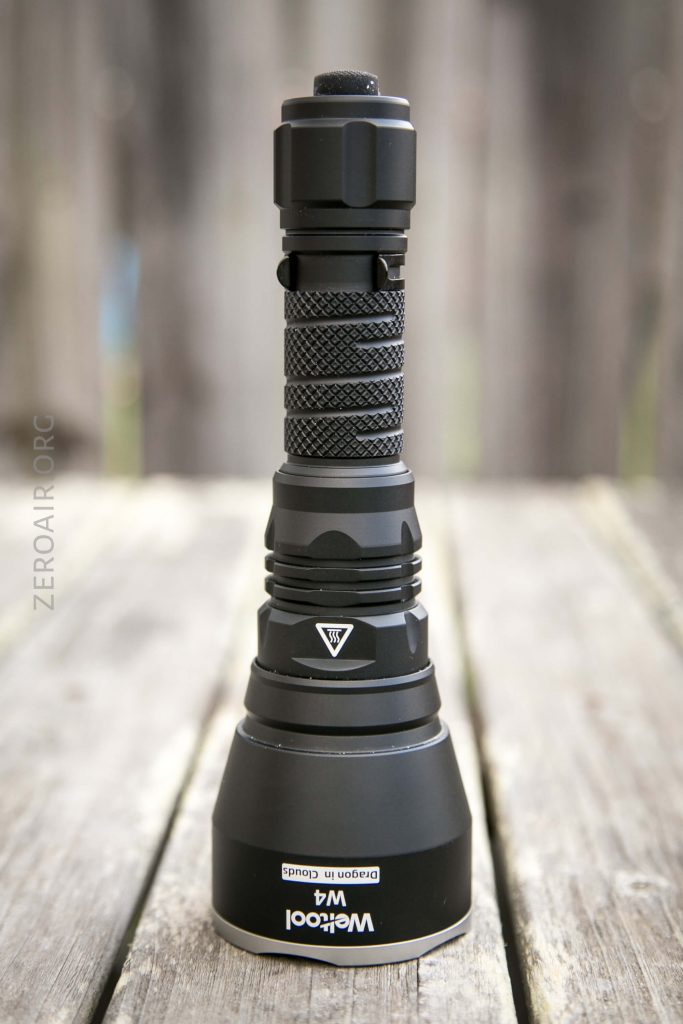 A black Weltool flashlight stands upright on a wooden surface with a blurred wooden background. The flashlight features a textured grip and a wide head section.