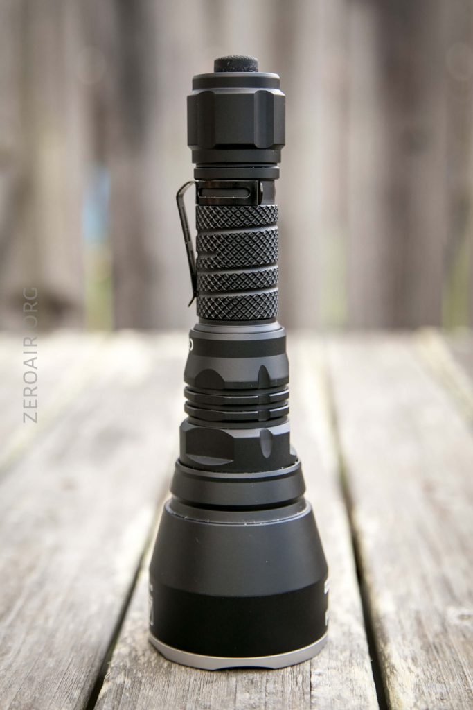 A black, handheld flashlight is standing upright on a wooden surface with a blurred wooden fence in the background. The flashlight has a textured grip and a wide head.