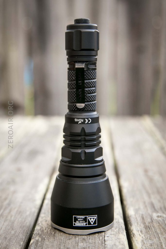 A black flashlight with a textured grip and clip stands upright on a wooden surface; a blurred wooden fence is in the background.