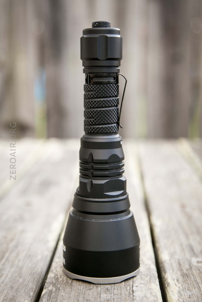 A black tactical flashlight with a textured grip and clip is standing upright on a wooden surface with a blurred background.