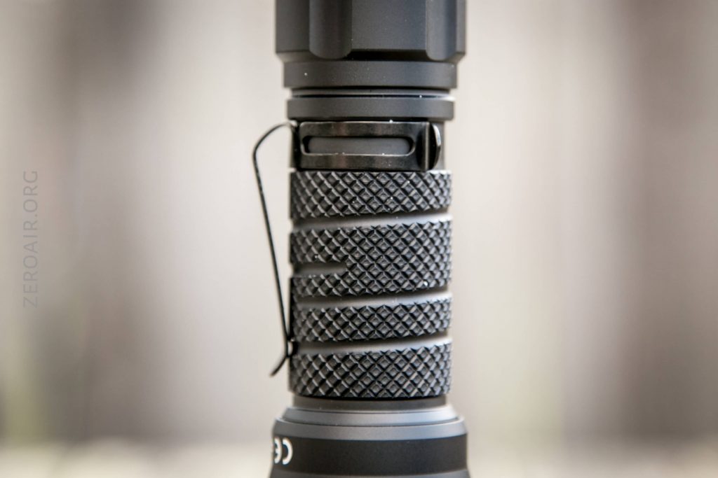 A close-up view of the textured grip and pocket clip on the body of a black flashlight. The image shows detailed knurling and part of the flashlight’s head and tail sections.