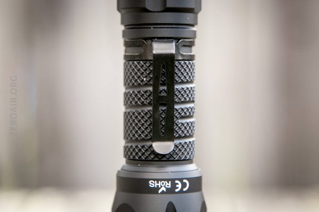 Close-up of a black flashlight handle showing a textured grip, a pocket clip, and certification markings including CE and RoHS on a matte black surface. Background is blurred.