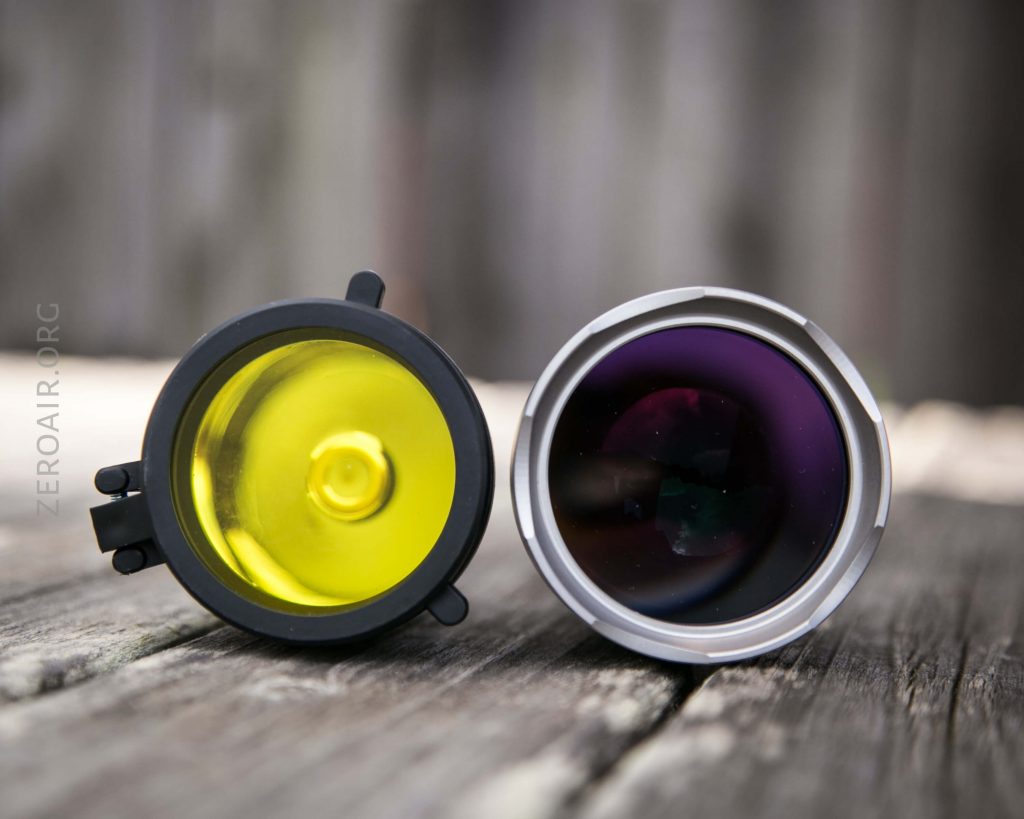 Two camera lenses rest side by side on a wooden surface; the left lens has a yellow tint, while the right lens has a clear, dark, reflective surface. The background is blurred.