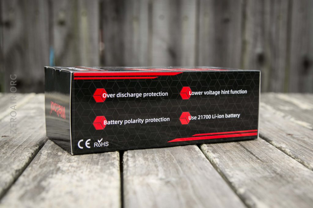 A product box sits on a wooden surface outdoors. Text on the box highlights features: over discharge protection, lower voltage hint function, battery polarity protection, and use of 21700 Li-ion battery. CE and RoHS marks are present.