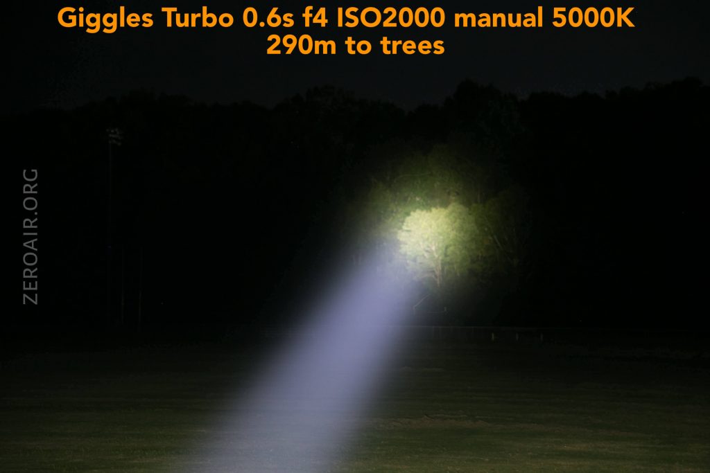 A bright, focused beam of light from a flashlight illuminates a patch of trees at night, with the camera settings and distance detailed in orange text at the top of the image.