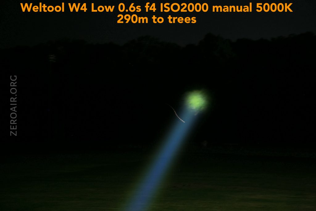 A bright blue flashlight beam aimed at a group of trees in the distance at night, with green light illuminating the treetops. Text above provides camera and flashlight settings used for the photo.