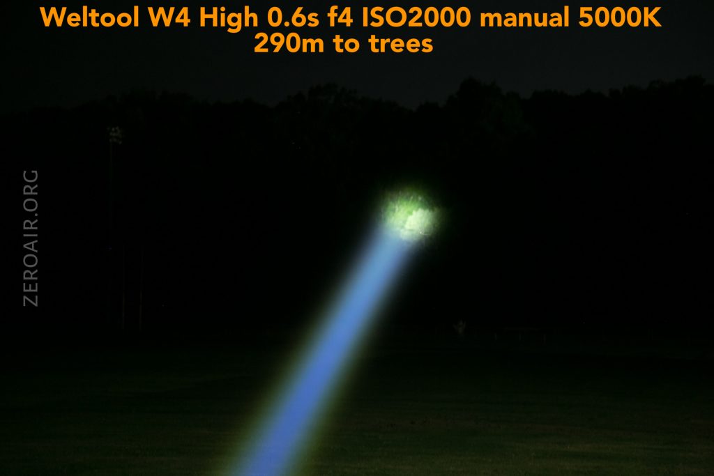 A bright flashlight beam extends from the foreground toward distant trees at night. Text at the top reads, Weltool W4 High 0.6s f4 ISO2000 manual 5000K 290m to trees. The background is mostly dark with faint tree outlines.
