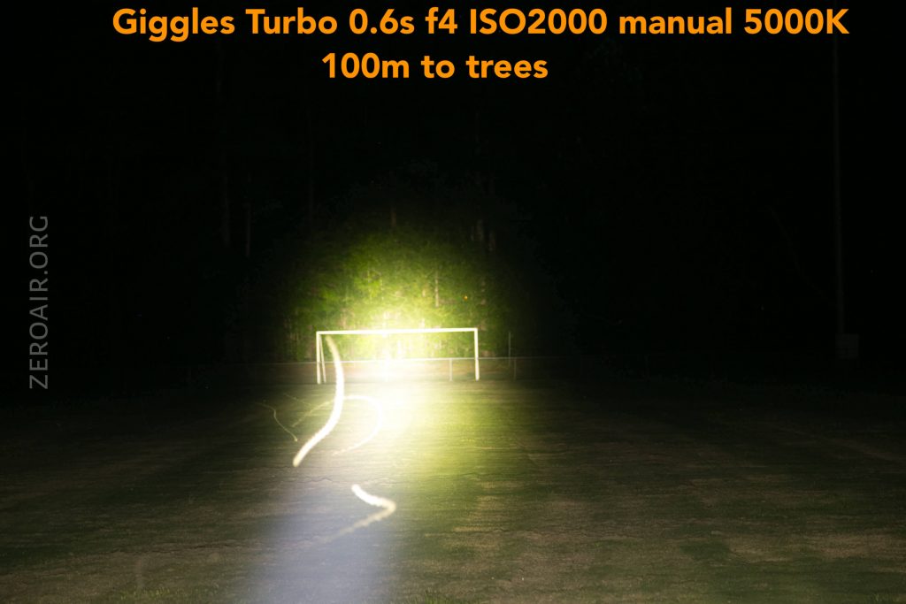 A bright flashlight beam illuminates a soccer goal in the distance at night, with camera settings and distance to trees noted in orange text at the top of the image. A light trail is visible on the grass.