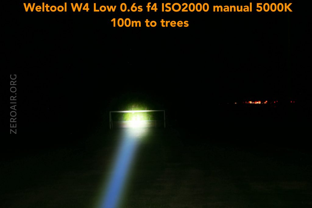 A bright flashlight beam shines on a distant soccer goal at night. The text at the top reads: “Weltool W4 Low 0.6s f4 ISO2000 manual 5000K 100m to trees.” The beam forms a visible path across the grass.