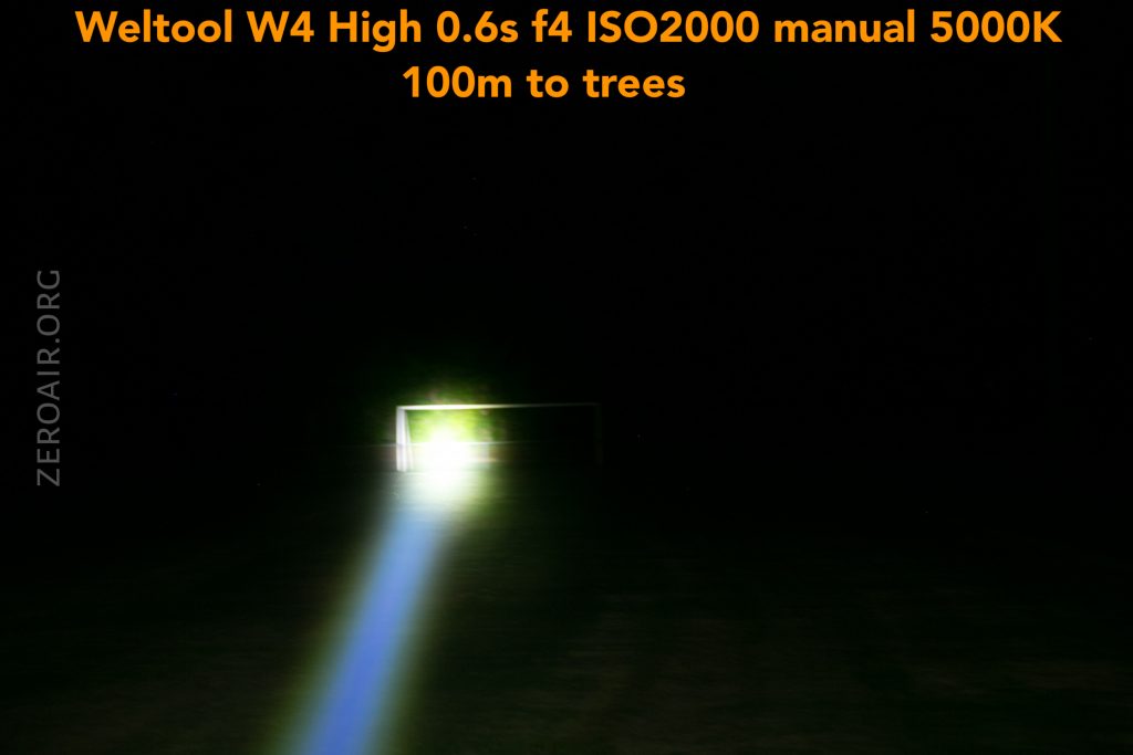 A bright flashlight beam shines directly ahead at a soccer goal on a dark field at night. Orange text at the top provides camera and flashlight settings, mentioning a 100-meter distance to the trees.