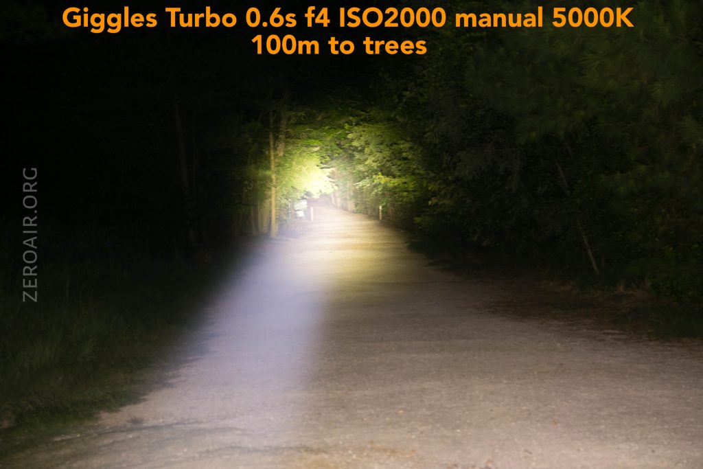A flashlight beam brightly illuminates a straight dirt road at night, reaching trees about 100 meters away. The surroundings are dim, with trees visible on both sides. Camera settings are displayed at the top.