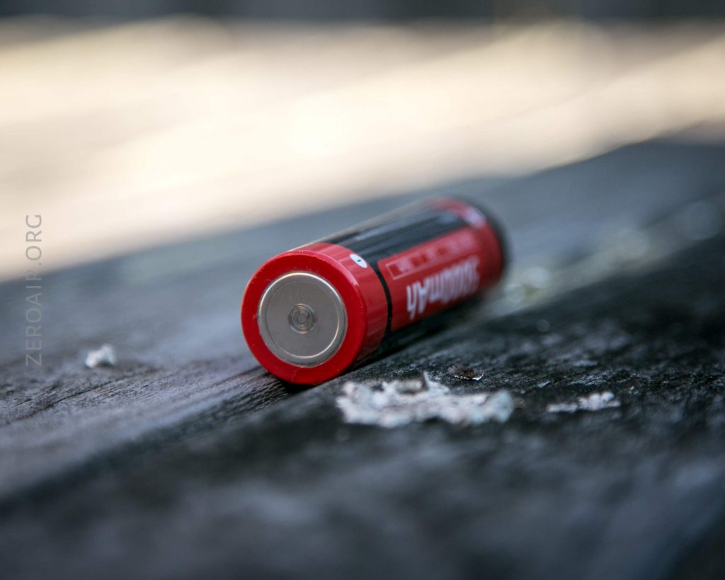 A single AA battery with a red and black casing lies on its side on a wooden surface. The battery’s positive terminal is facing the camera, and there are small wood shavings nearby.