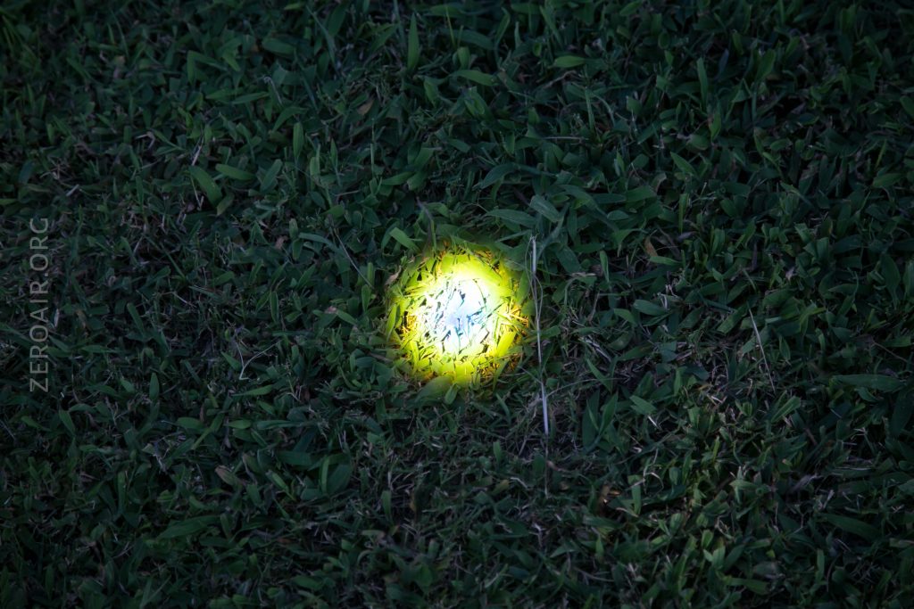A patch of green grass is illuminated by a circular beam of bright yellowish light, highlighting the blades in the center while the surrounding grass remains in shadow.