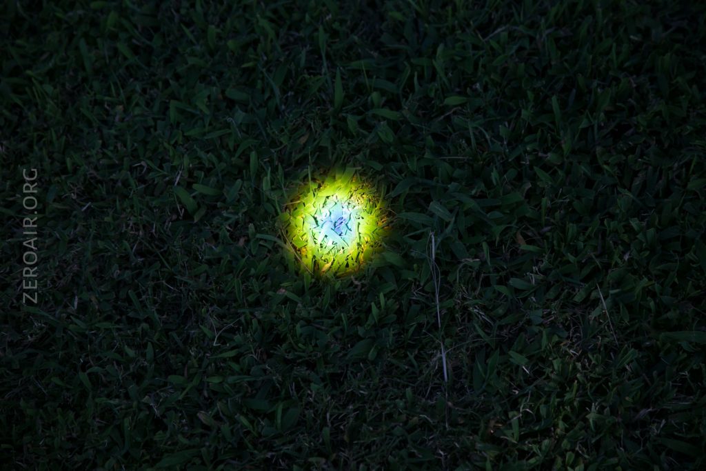 A small, bright yellow and white circular light shines on dark green grass at night, illuminating a patch in the center of the image. The background is mostly obscured by shadows. The text ZEROAIR.ORG appears on the left side.