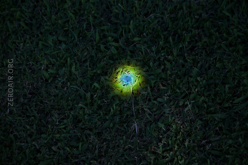 A circular patch of grass illuminated by a small, bright beam of light with blue and yellow hues; the surrounding grass remains dark. The website “ZEROAIR.ORG” appears along the left side of the image.