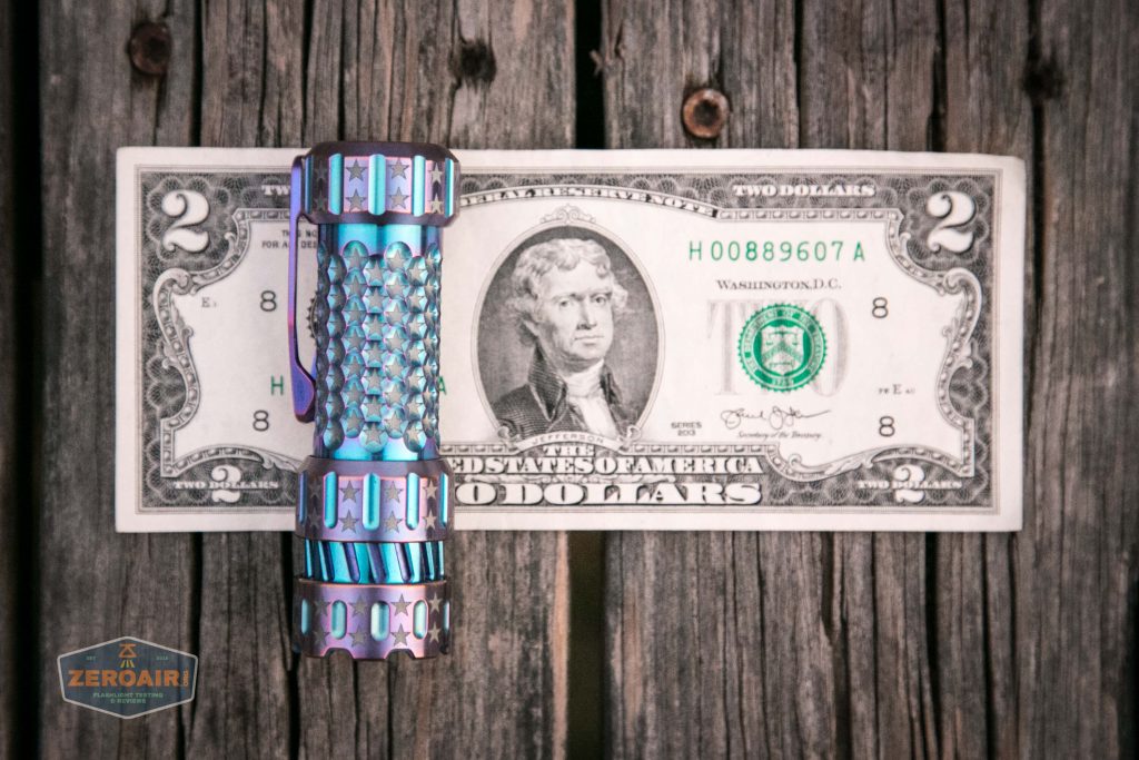 A metallic, patterned flashlight rests horizontally in front of a United States two-dollar bill, both placed on a wooden surface with visible grain and screws.