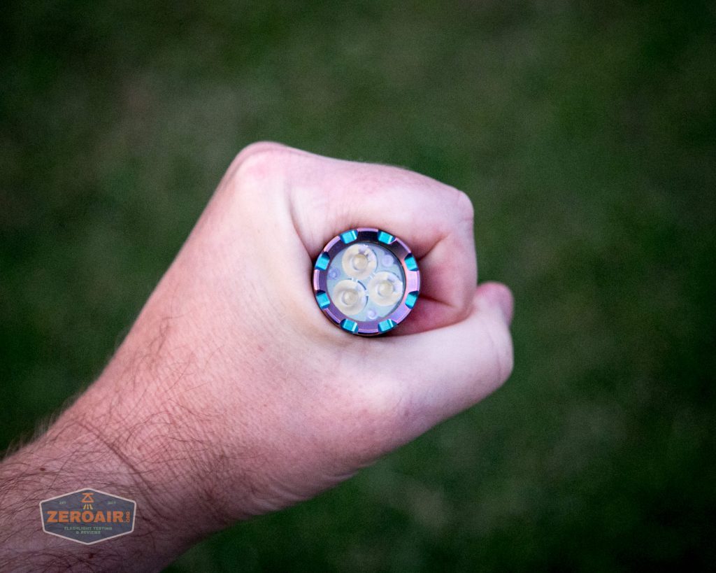 A person’s hand holding a small flashlight facing the camera, showing four LED bulbs arranged in a circular pattern against a blurred green background.