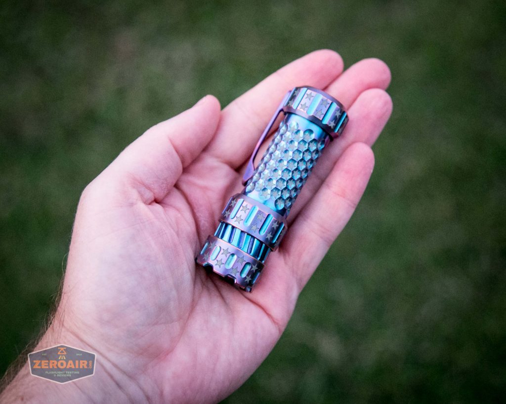 A hand holding a small, metallic flashlight with a textured grip and iridescent blue and purple coloring. The background is out-of-focus grass. There is a ZeroAir watermark in the lower left corner.