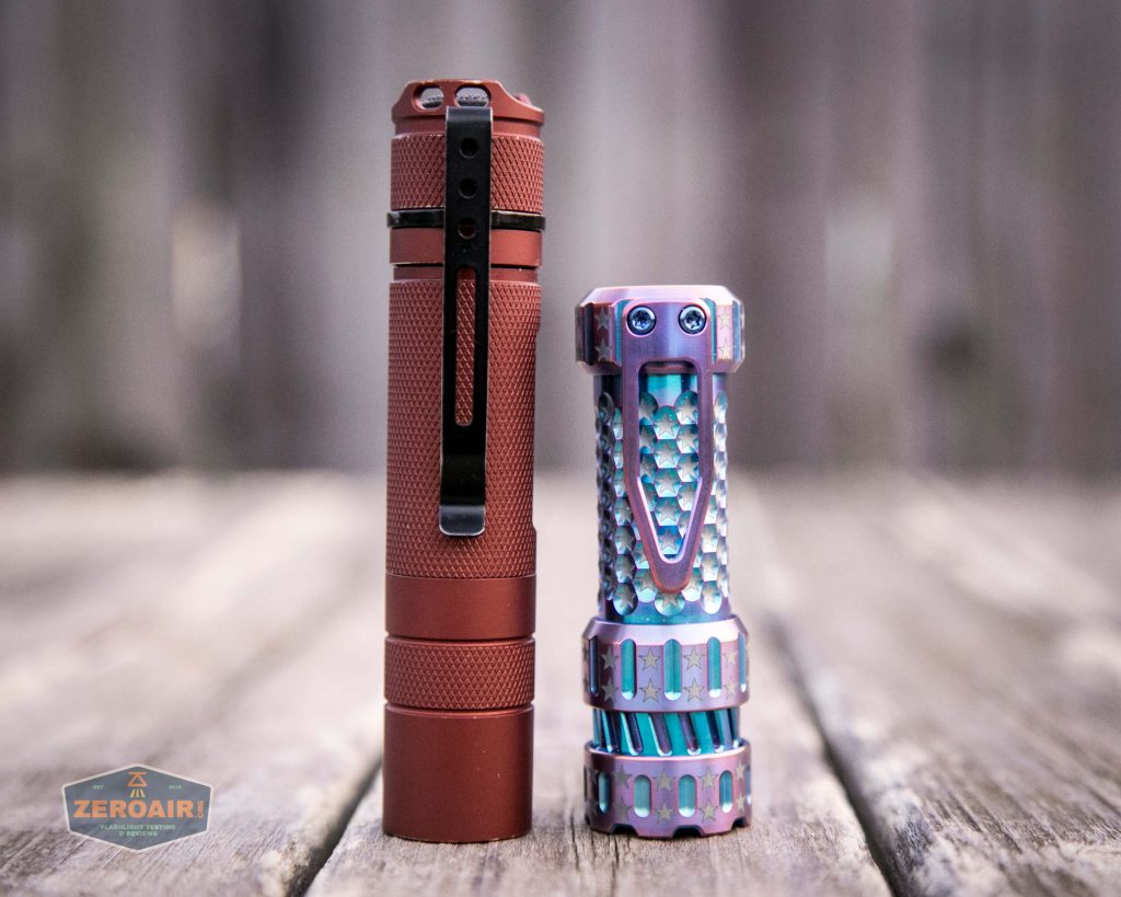 Two compact flashlights stand upright on a wooden surface. The left flashlight is red with a black pocket clip, while the right flashlight is metallic with a textured, multi-colored pattern. The background is blurred.