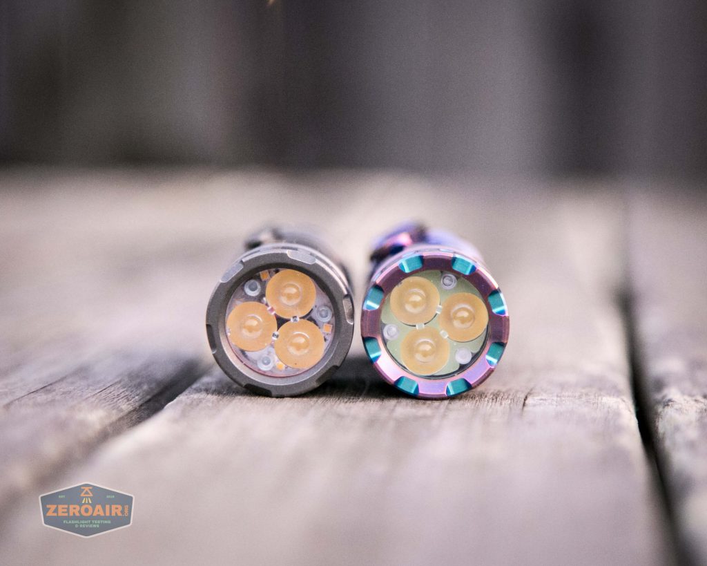 Two small, cylindrical flashlights are placed side by side on a wooden surface, with their LED emitters facing the camera. The left flashlight is silver, and the right one has a multicolored finish.