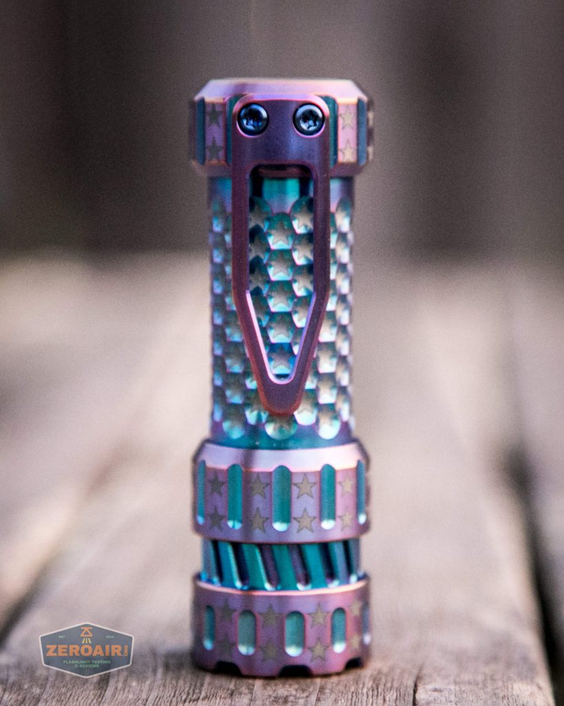 A metallic, textured flashlight with a patterned grip and a large clip is standing upright on a wooden surface. The flashlight has a pink and blue finish with star designs. A logo reading ZeroAir! is in the corner.
