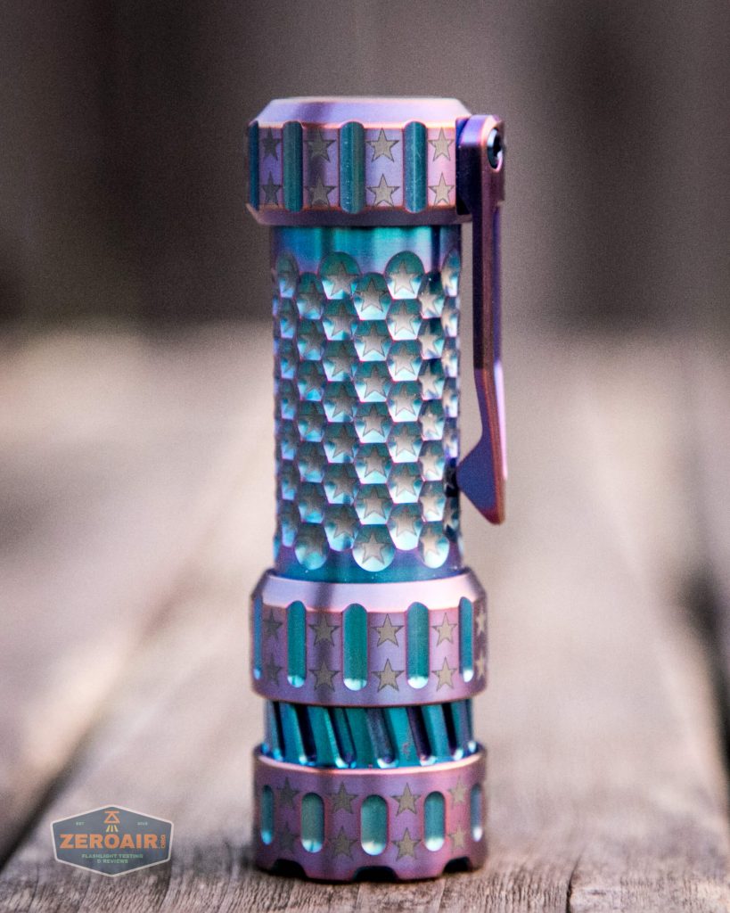 A small, metallic flashlight with a textured honeycomb pattern, star engravings, and a pocket clip stands upright on a wooden surface. The finish appears iridescent with shades of blue and purple.