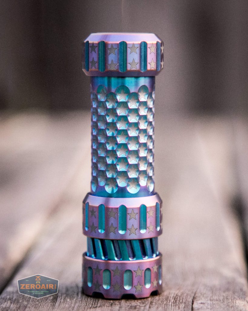 A metallic, cylindrical flashlight with a textured honeycomb grip and star-shaped engravings stands upright on a wooden surface. The flashlight has a blue and purple gradient finish.