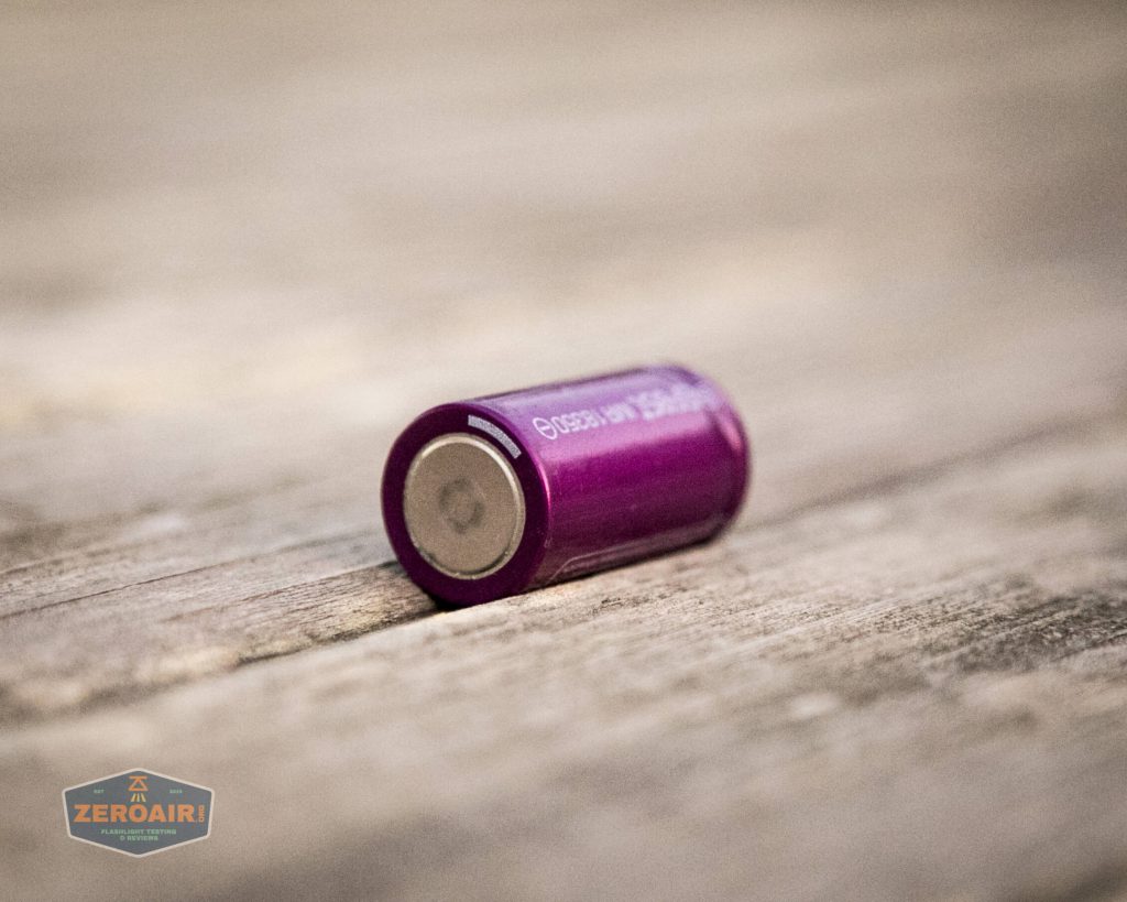A single purple cylindrical battery lying on its side on a weathered wooden surface. The batterys positive terminal is visible. A blurred logo reading ZEROAIR appears in the lower left corner.
