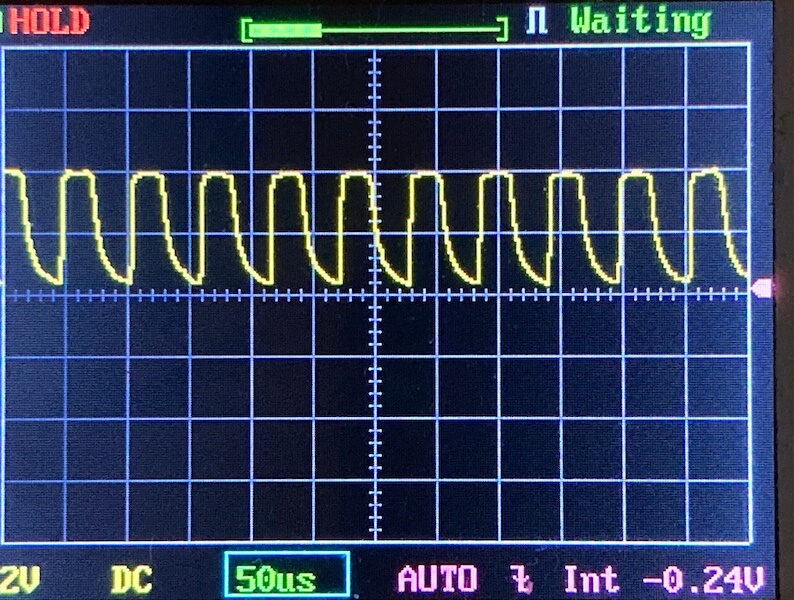 An oscilloscope screen displays a repeating yellow waveform with irregular peaks and troughs. The time base is set to 50 microseconds per division. The display shows voltage, DC mode, and other measurement information.