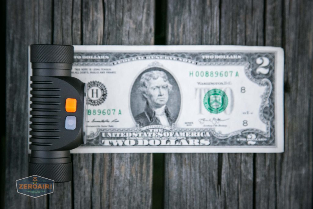 A small black flashlight is placed next to a two-dollar bill on a wooden surface, showing their size comparison. The flashlight has two buttons, one orange and one gray.