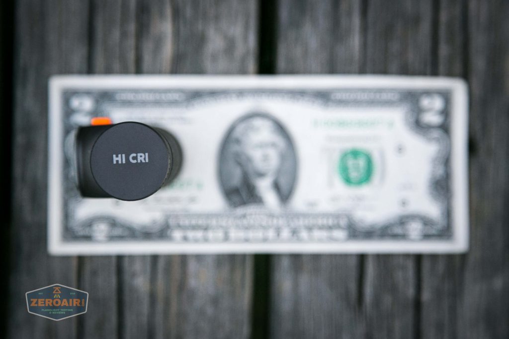 A round flashlight head labeled HI CRI is positioned vertically on top of a blurred U.S. two-dollar bill, resting on a wooden surface. The ZeroAir logo appears in the lower left corner.
