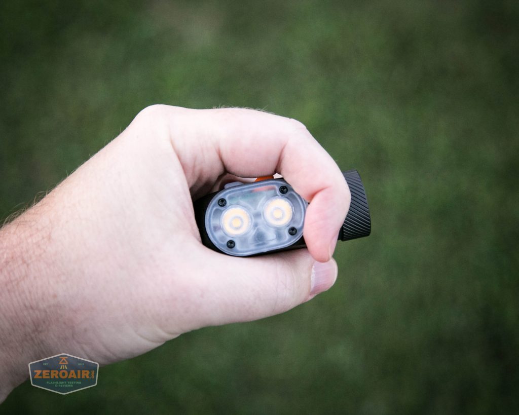 A left hand holding a black rectangular flashlight with two visible LED bulbs, positioned above a grassy surface outdoors. The ZeroAir watermark appears in the bottom left corner.