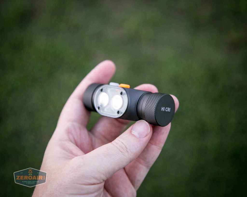 A hand holding a compact black flashlight with two LED bulbs and HI CRI written on one end, shown against a blurry outdoor green background.