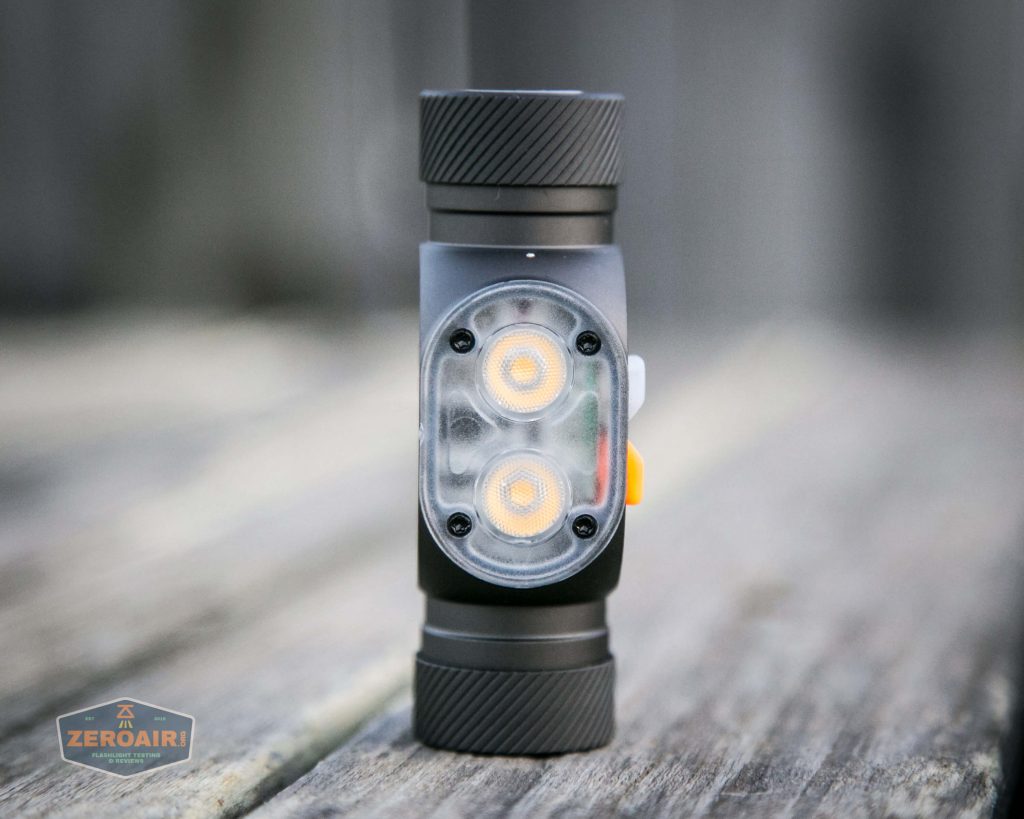 A compact, black flashlight with two circular LED lights on the front stands upright on a wooden surface. The flashlight has textured ends and a visible orange accent button on the side.