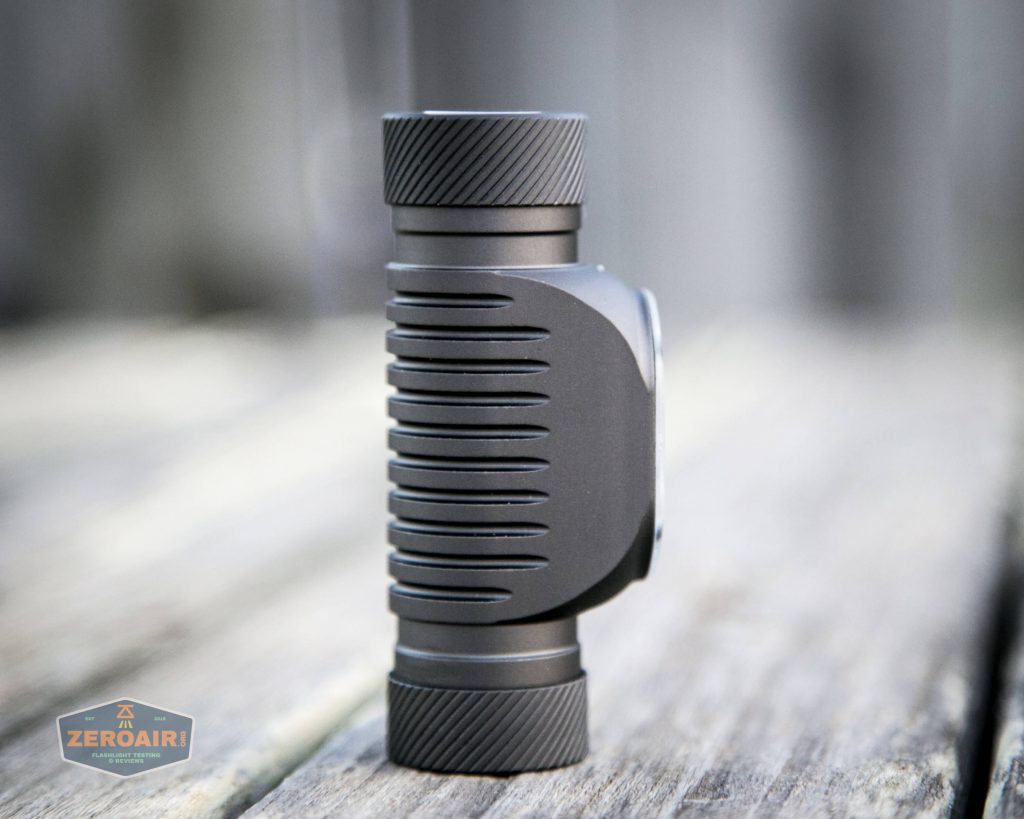 A compact black cylindrical flashlight with ribbed sides and textured end caps stands upright on a wooden surface. The background is blurred. A small ZeroAir logo is visible in the lower left corner.