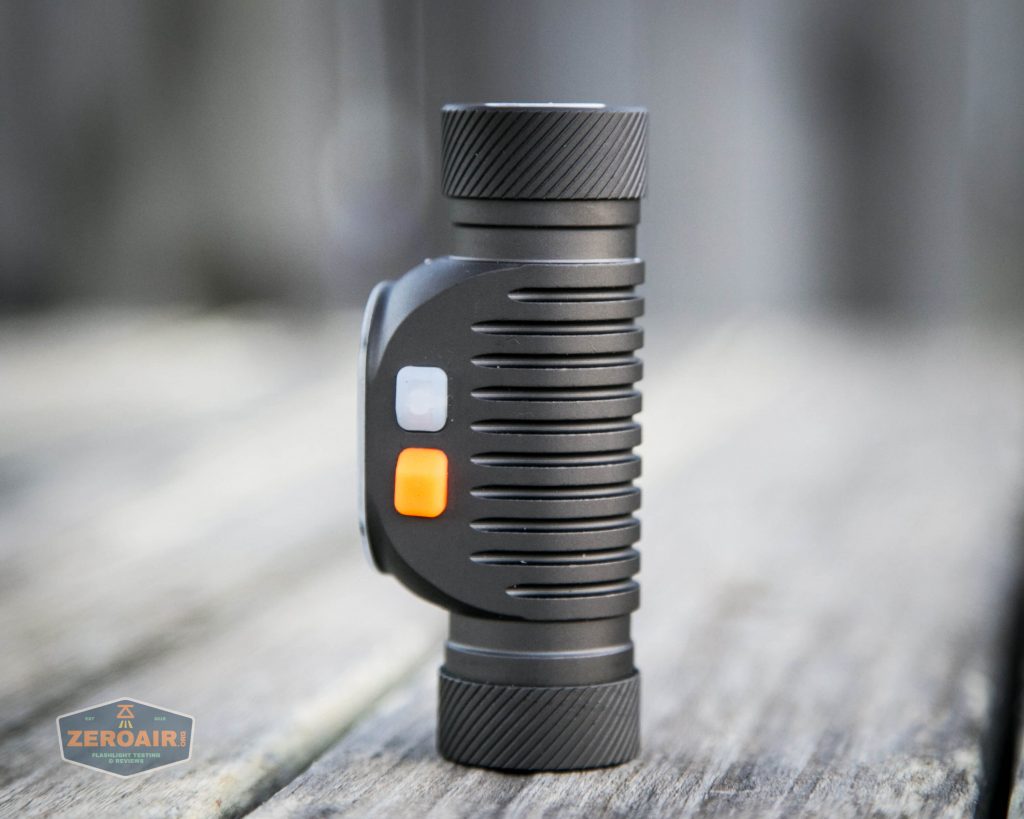 A compact, black, cylindrical flashlight with ribbed sides stands upright on a wooden surface. It has two square buttons—one white and one orange—on its side. A ZeroAir logo is visible in the bottom left corner.