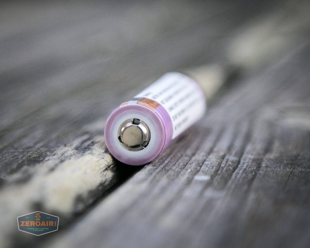 A single cylindrical battery with a purple ring around the positive terminal lies on a weathered wooden surface. The battery is partially out of focus, and a ZeroAir watermark appears in the bottom left corner.