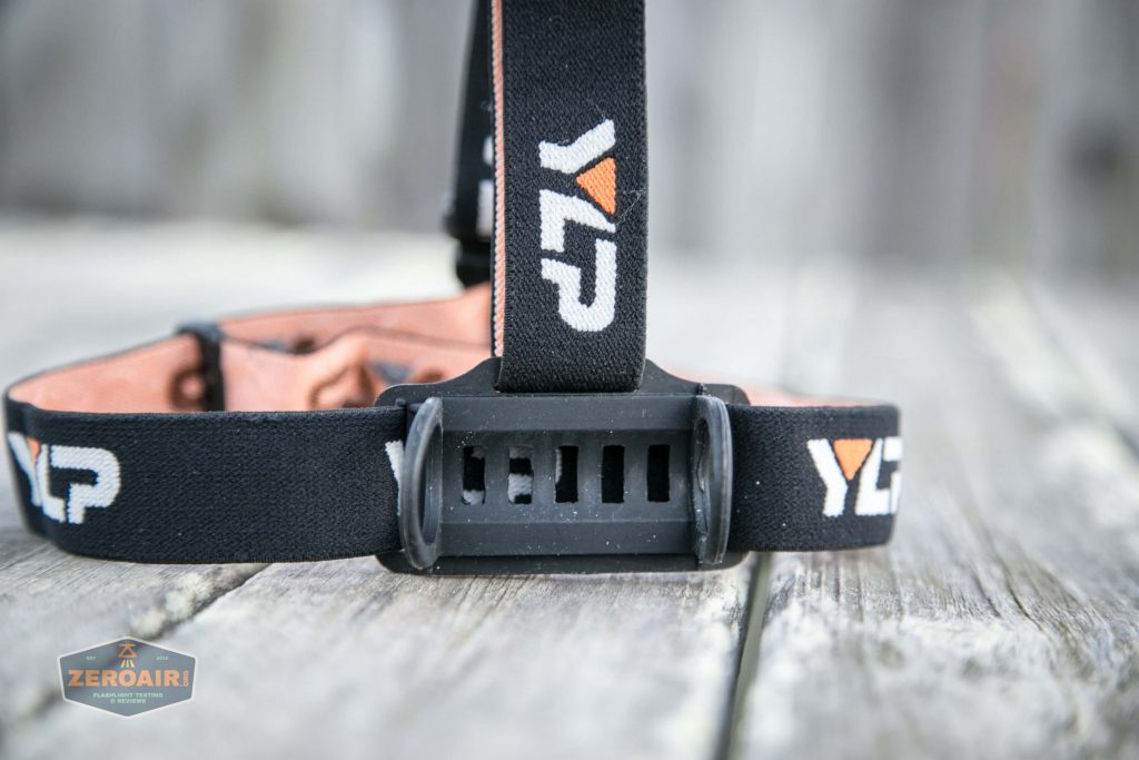 A close-up of a black and orange headlamp strap with the letters YLP printed on the band. The strap is attached to a plastic mount and is resting on a weathered wooden surface.