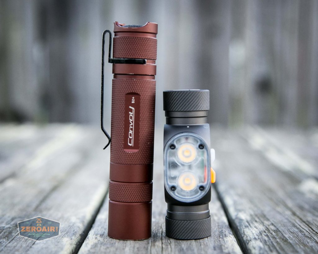 Two flashlights stand upright on a weathered wooden surface. The flashlight on the left is slim and red with a metal clip, while the one on the right is shorter, gray, and has two visible LEDs. A blurred background is visible.